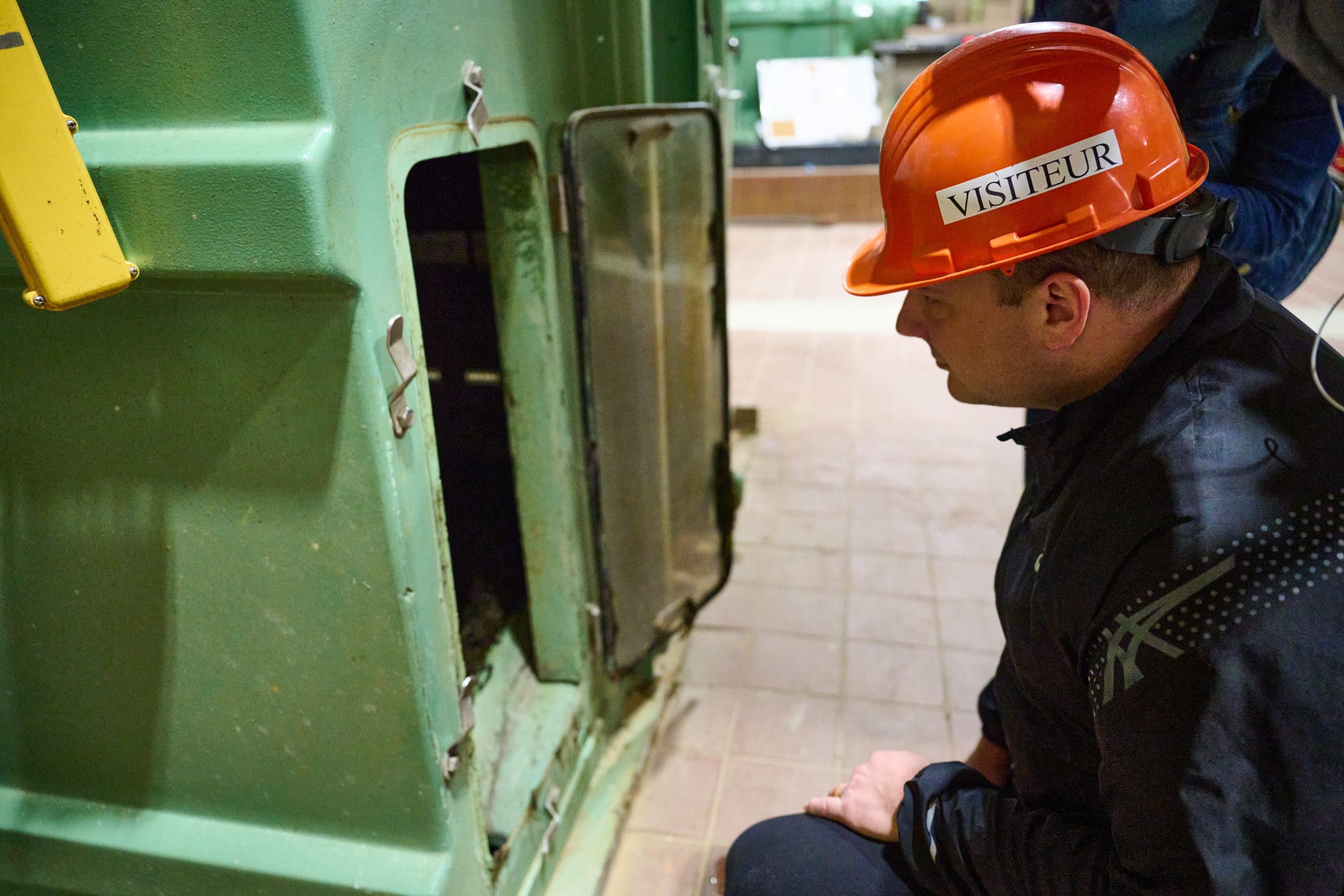 A man with an orange safety helmet inspects a green machine with a service door opened.