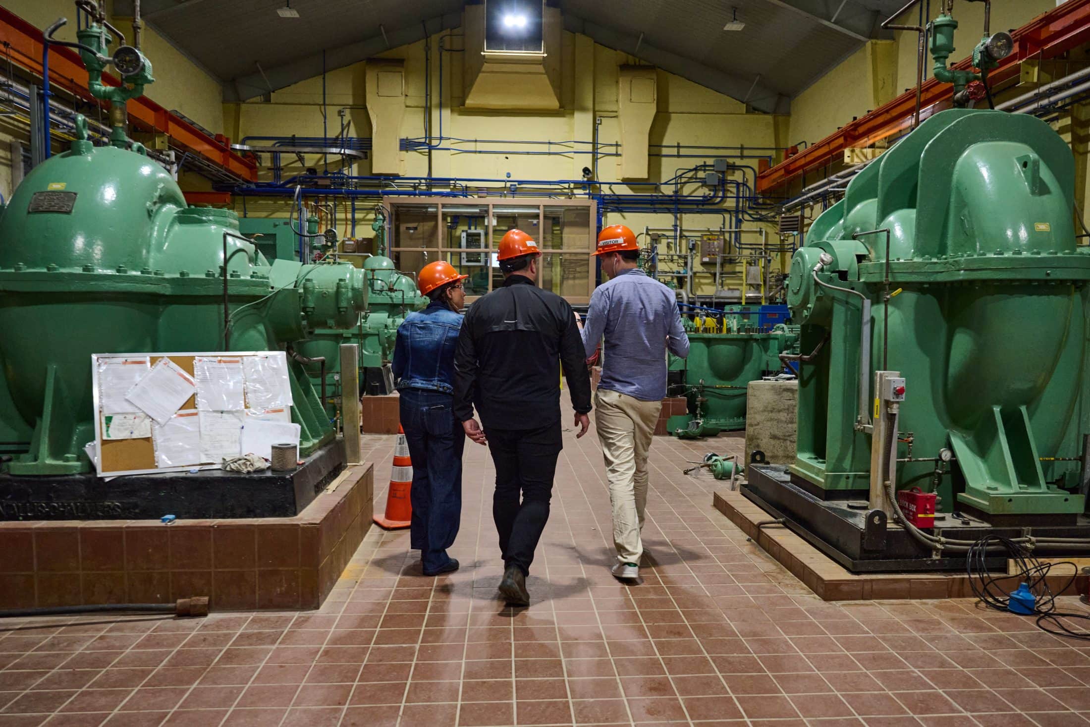 Three people with orange safety helmets walk through an industrial room with large green pumps.