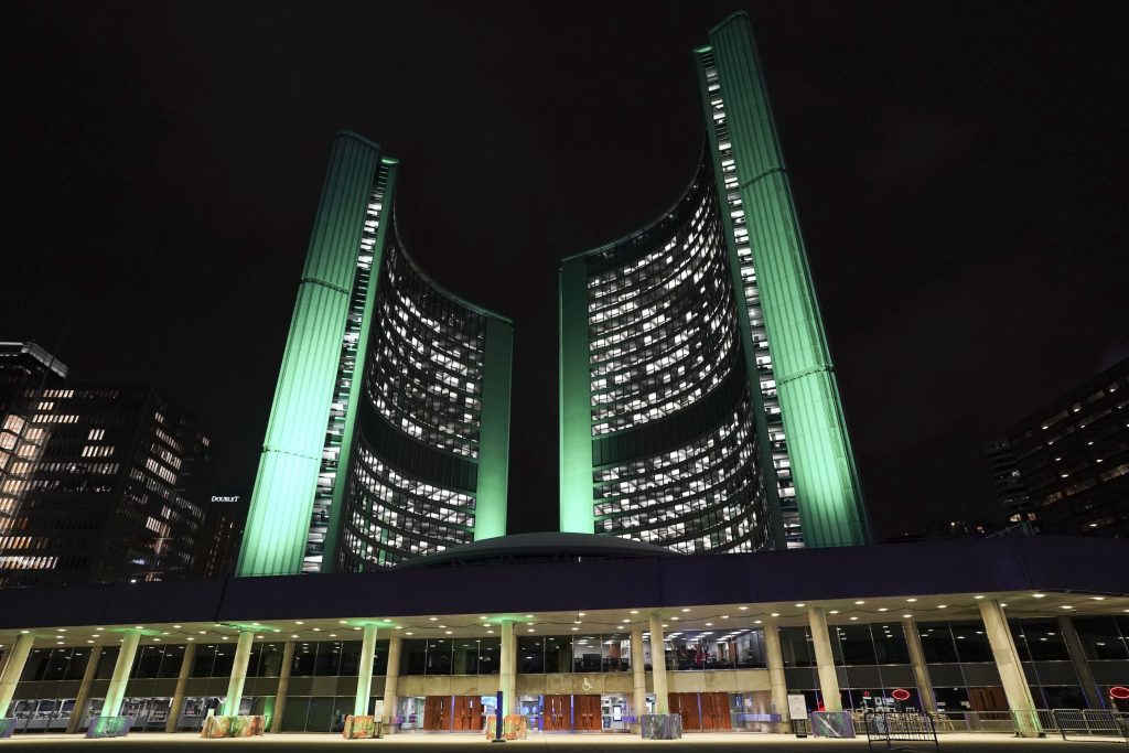 Toronto City Hall is illuminated at night, with green lights highlighting the curved towers rising above the council chambers.