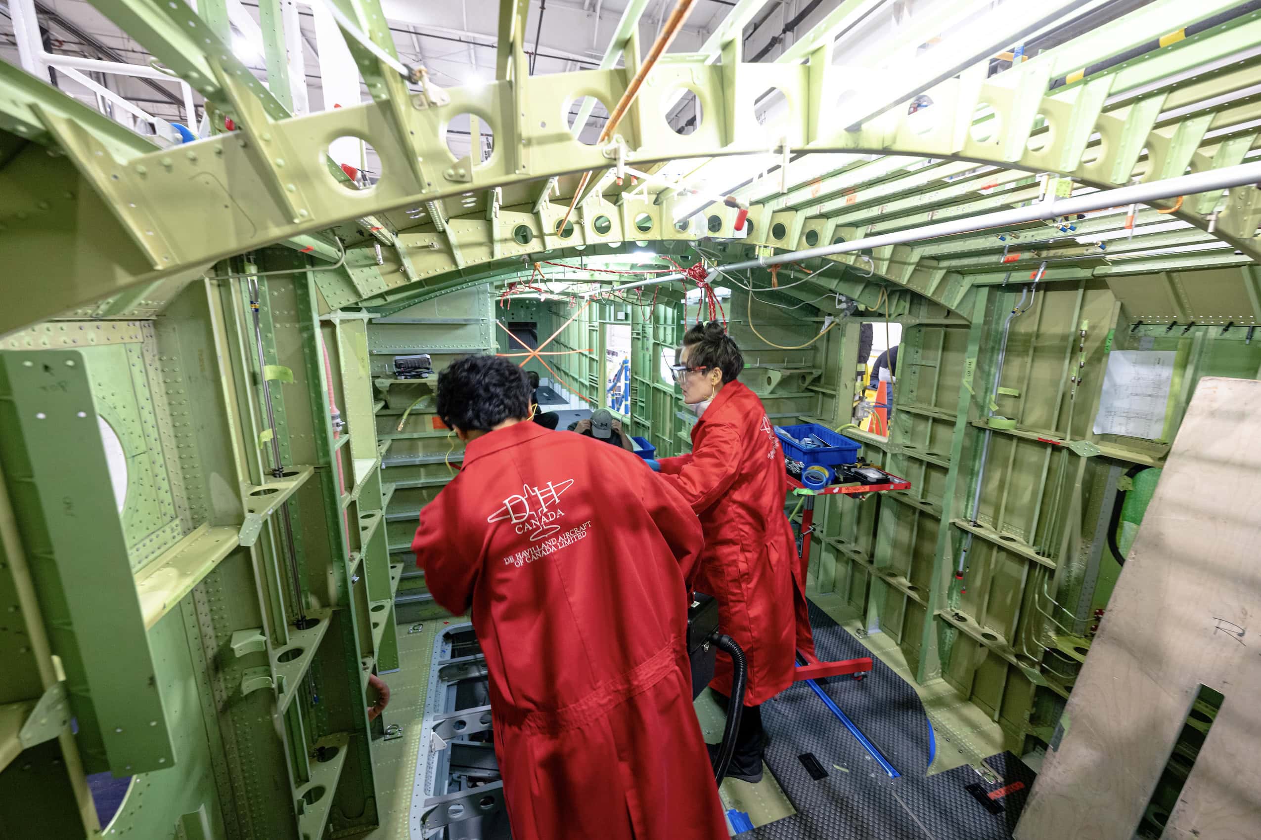 Two workers wearing red coats are inside an unfinished water bomber aircraft during its construction.